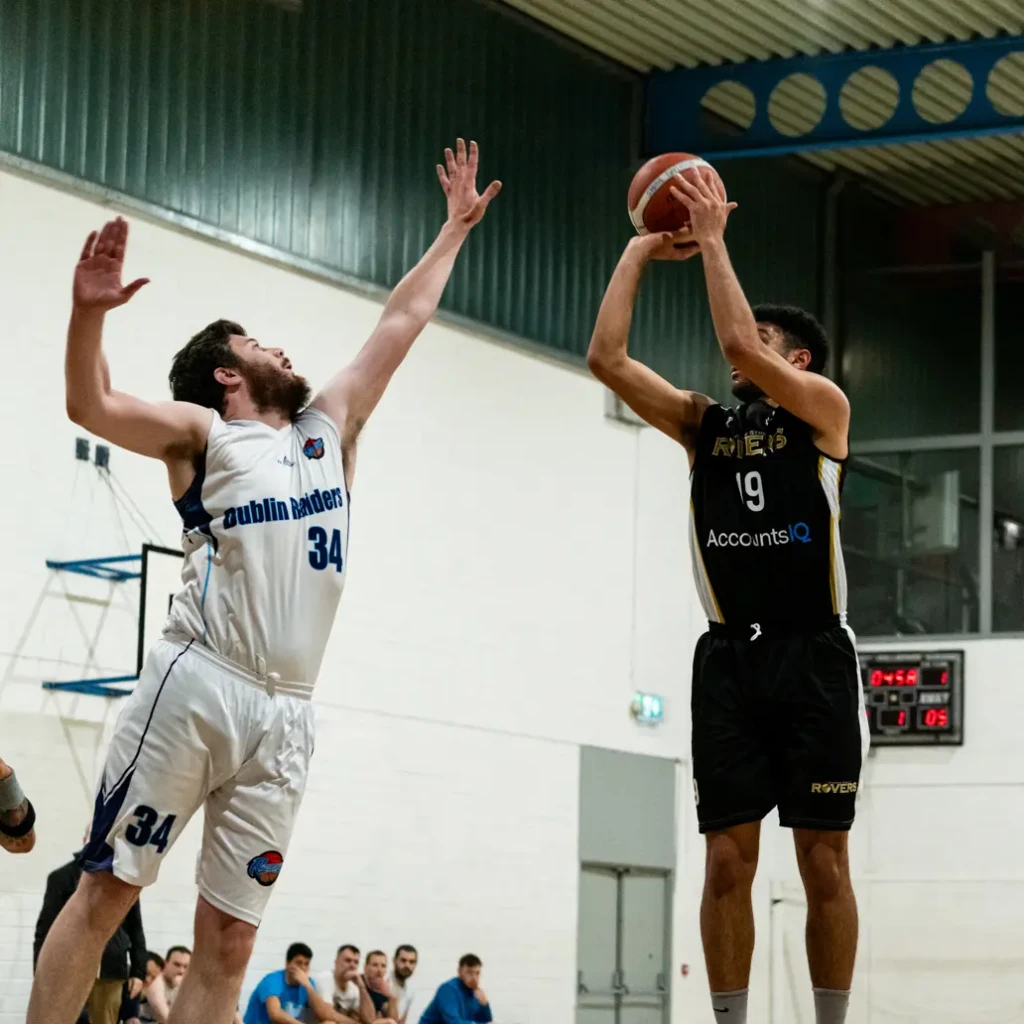 Joueur de basketball en tir en suspension face à un défenseur bras levé lors d'un match en salle